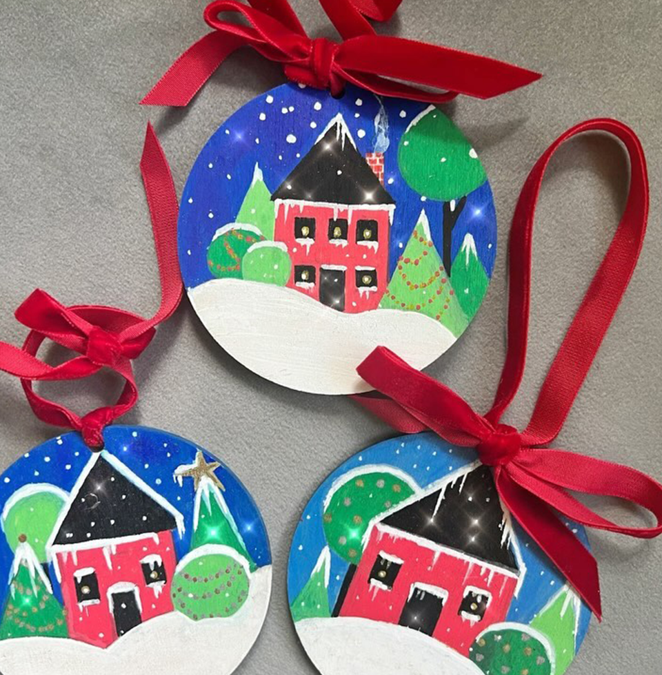 Three circular Christmas ornaments with red ribbons, featuring hand-painted winter scenes of red houses, snow, and trees against a blue sky.