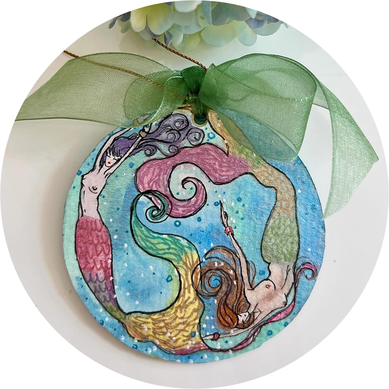 A round ornament features three colorful mermaids with flowing hair, arranged in a circular pattern. A green ribbon is tied at the top for hanging.